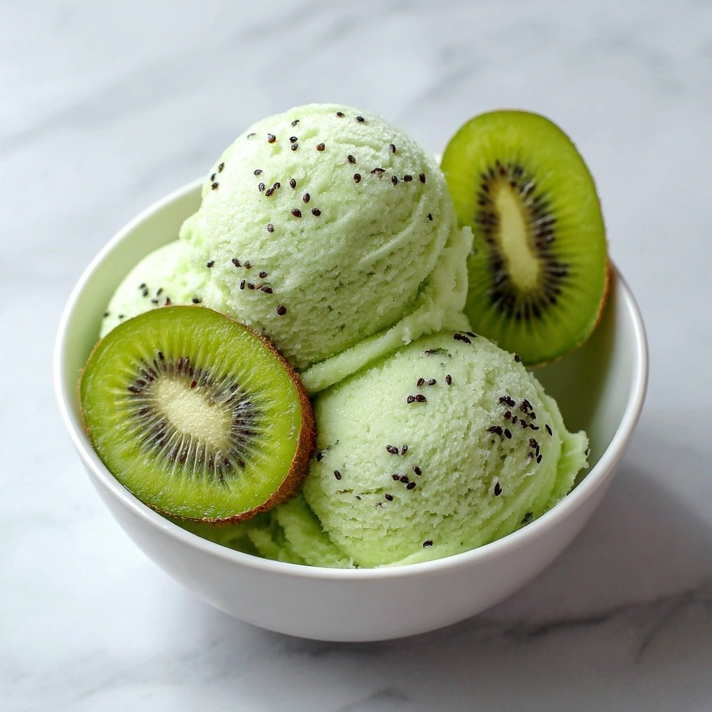 The image shows a white bowl filled with three scoops of pale green kiwi sorbet, each scoop sprinkled with small black kiwi seeds for texture. There are fresh green kiwi slices placed inside the bowl against the sorbet, showing the bright green flesh and radiating white core with tiny black seeds. The bowl rests on a white marbled surface, and the scene is softly lit, highlighting the smooth, icy texture of the sorbet and the freshness of the kiwi slices. photo taken with an iphone --ar 1:1 --v 7 — Kiwi Sorbet, kiwi dessert, healthy sorbet recipe, vegan frozen treat, easy fruit sorbet